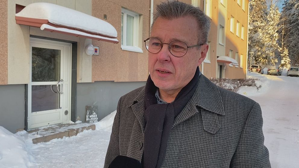 En man klädd i en mörk kostymjacka står utomhus framför en flerfamiljshus täckt av snö. I bakgrunden syns en entrédörr och snötäckta bilar längs gatan som skapar en vinterlig atmosfär.