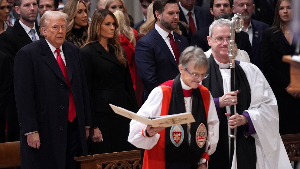 Prästen Marianne Edgar Budde klädd i en röd och svart kappa står framåtlutad och läser ur en bok, medan flera personer i formell klädsel står bakom. USA:s president Donald Trump står till vänster, med en röd slips. Han  är uppenbart framträdande i samlingen. Atmosfären är högtidlig och samlad.