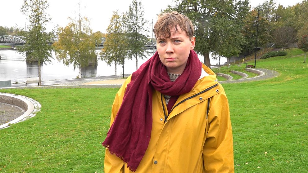 Ellen Ström står på en grön gräsmatta med en gul regnjacka och en röd scarf. I bakgrunden syns Umeåälven med spegelblanka ytor och omgivande träd, vilket ger en höstlig atmosfär. Det är en lugn och svagt molnig dag.