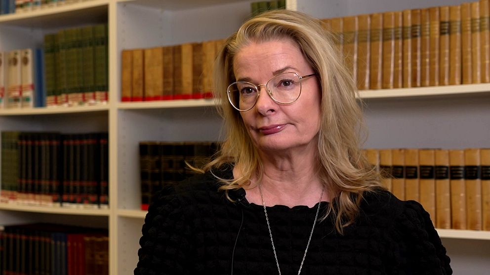 En kvinna med axellångt blont hår står framför en bokhylla fylld med inbundna böcker.