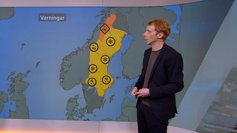 En person står framför en väderkarta som visar varningar för olika väderförhållanden i Sverige. Kartan har områden markerade i orange och gult som indikerar varningar, tillsammans med symboler för snö och andra möjliga faror. Texten ”Varningar” är synlig i övre vänstra hörnet av kartan.