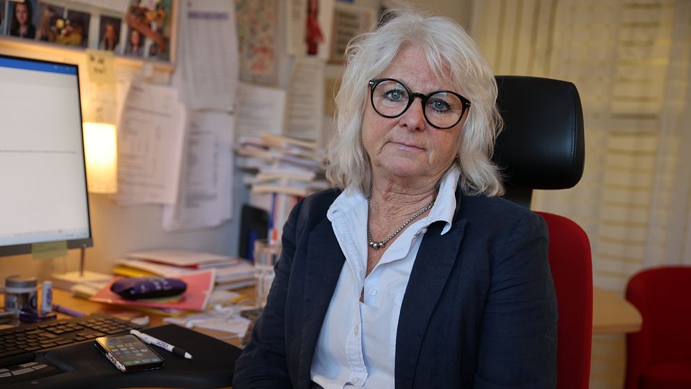 En person sitter vid ett skrivbord i ett kontorslandskap, omgiven av högar av papper och kontorsmaterial. På skrivbordet syns en dator med en öppen skärm och diverse föremål som en mobiltelefon och en penna. Bakgrunden är fylld med dokument och notiser som hänger på väggen.