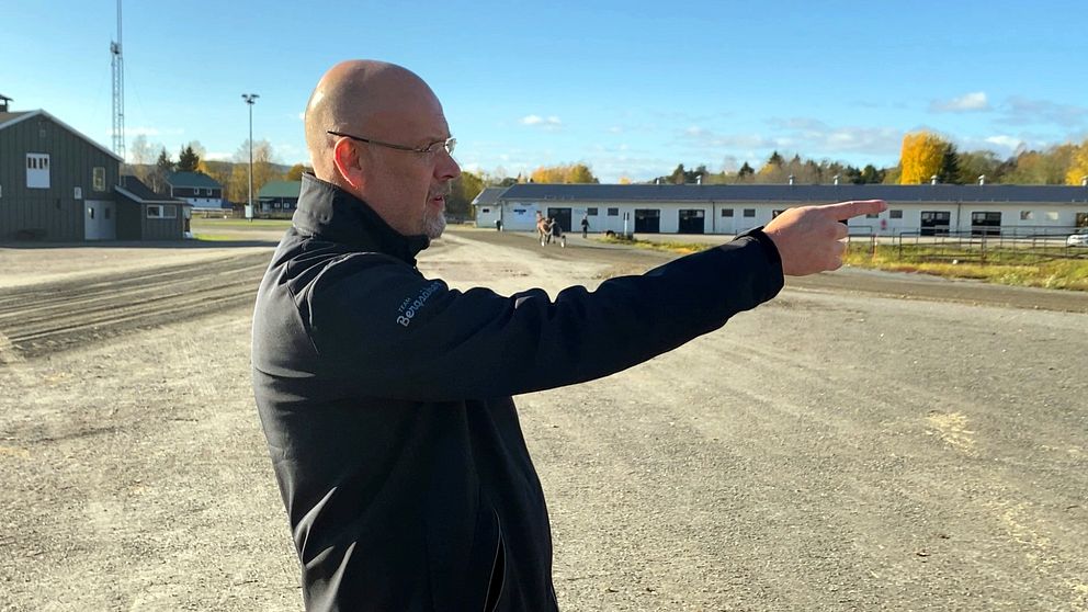 En man står på en grusplan och pekar framåt med höger hand. I bakgrunden syns byggnader och en travghäst med ett höstigt landskap. Himlen är klar med några få moln.