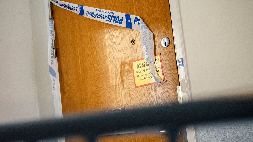 En stängd dörr märkt med polisens avspärrningsband och en gul skylt med texten ”AVSPÄRRAT”. Dörren visar tecken på slitage med fläckar synliga på ytan. Misstänkt mord i Lund.