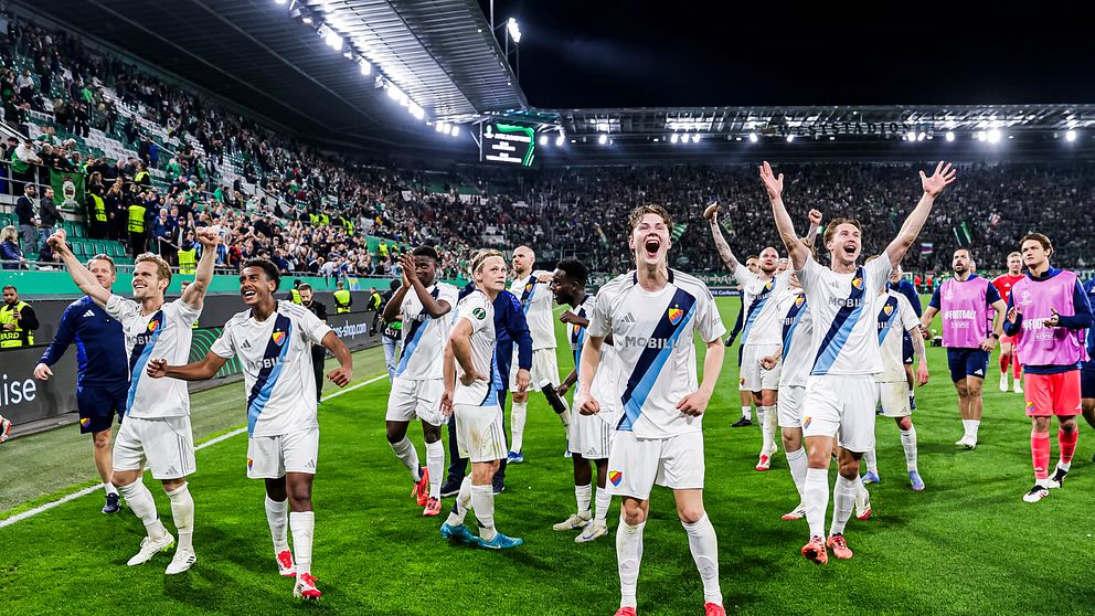 Fotbollsspelare i vita matchställ jublar framför publik på fullsatt arena.