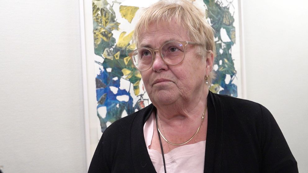 En äldre kvinna med ljust hår står framför en abstrakt tavla på en vit vägg.