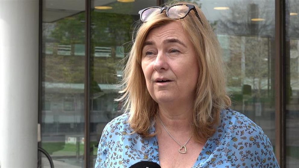 Kvinna med blont hår och blåmönstrad skjorta intervjuas utomhus framför glasdörrar.