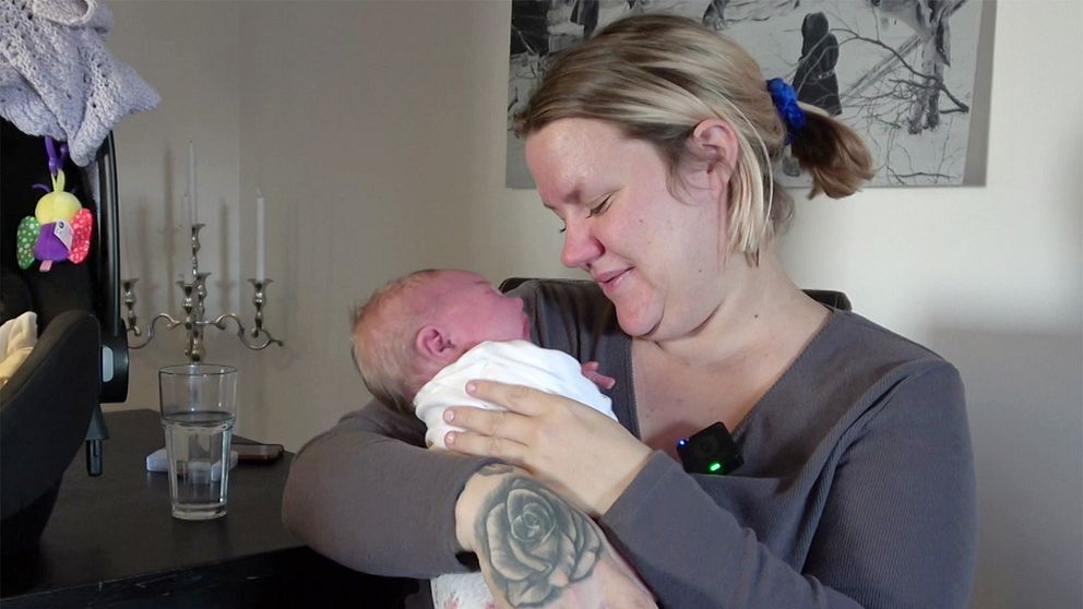 Åsa Asplund håller sitt nyfödda barn i sina armar, med en mjuk och kärleksfull gest. I bakgrunden syns ljusare inredning och ett par ljusstakar, vilket bidrar till en hemtrevlig atmosfär. Ett glas vatten står på bordet bredvid.