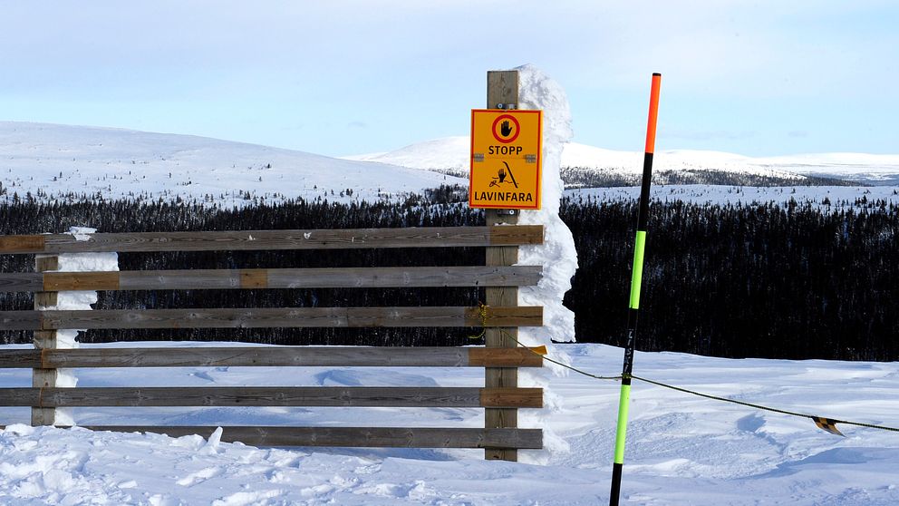 Skylt varning för lavinrisk vid snötäckt berg med trägrind och skidstolpe.