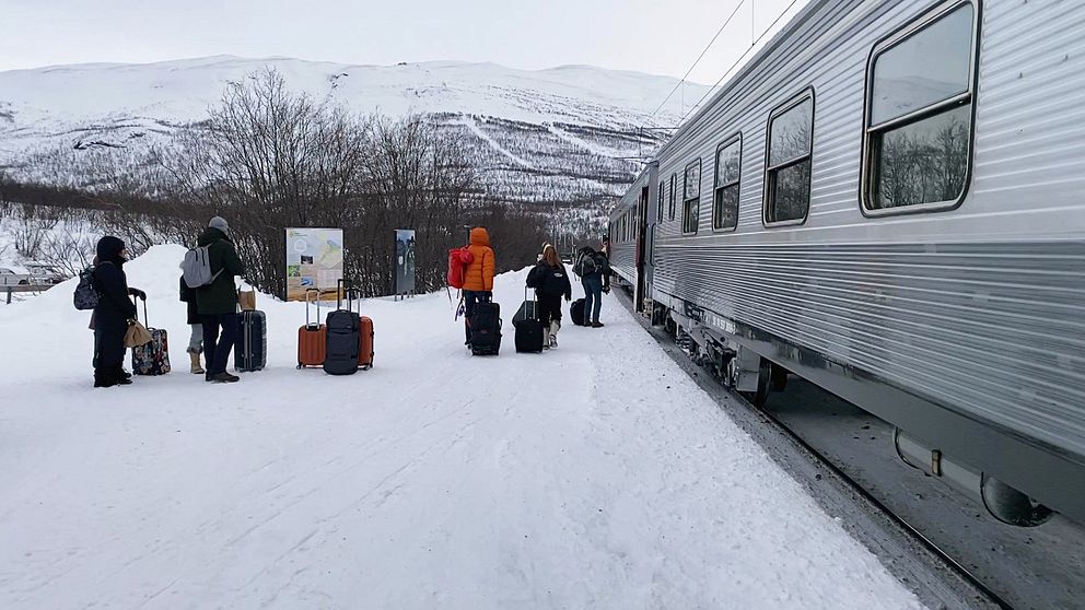 Passagerare väntar på en tågstation i ett snöigt landskap, med en tågvagn synlig till höger. Flera personer är klädda i vinterkläder och har bagage. I bakgrunden syns snötäckta fjäll och en informationsskylt vid stationen.