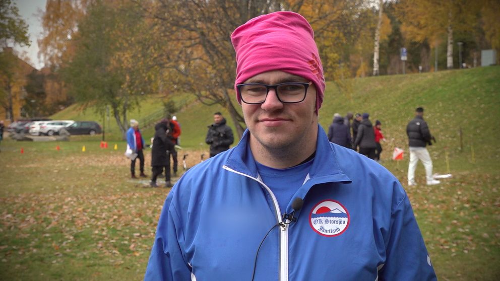 Henrik Berglund, från orienteringsklubben OK Storsjön, står utomhus med ljusblå klubbjacka och rosa mössa.