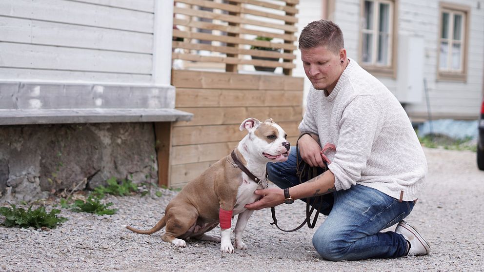 Man sitter på huk utomhus och håller i en hund med koppel nära ett hus.