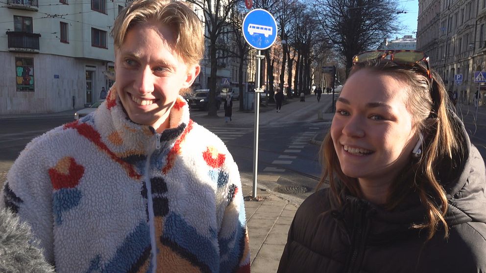 Två personer står på en trottoar i en stadsmiljö. En blå skylt, som indikerar en busshållplats, är synlig i bakgrunden. En bred gata kantad av byggnader och träd sträcker sig bortom personerna. Det är en solig dag.