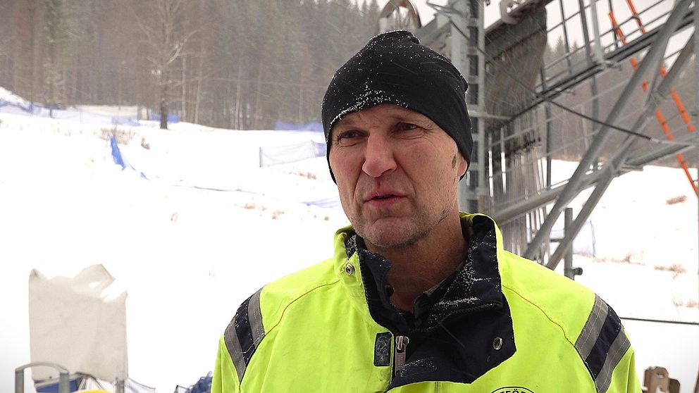 Mattias Wikner står i en gul arbetsjacka i en snöig miljö, framför liftanläggningen i Persåsen med snötäckta träd i bakgrunden.