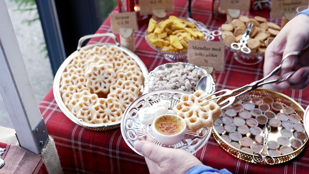 En hand plockar upp en krispig kaka från en silverfärgad tallrik, där det även finns en liten skål med en krämig dip. I bakgrunden syns flera olika sorters godsaker som är vackert presenterade på ett rödvitrutigt bord. Små skyltar beskriver de olika delikatesserna.