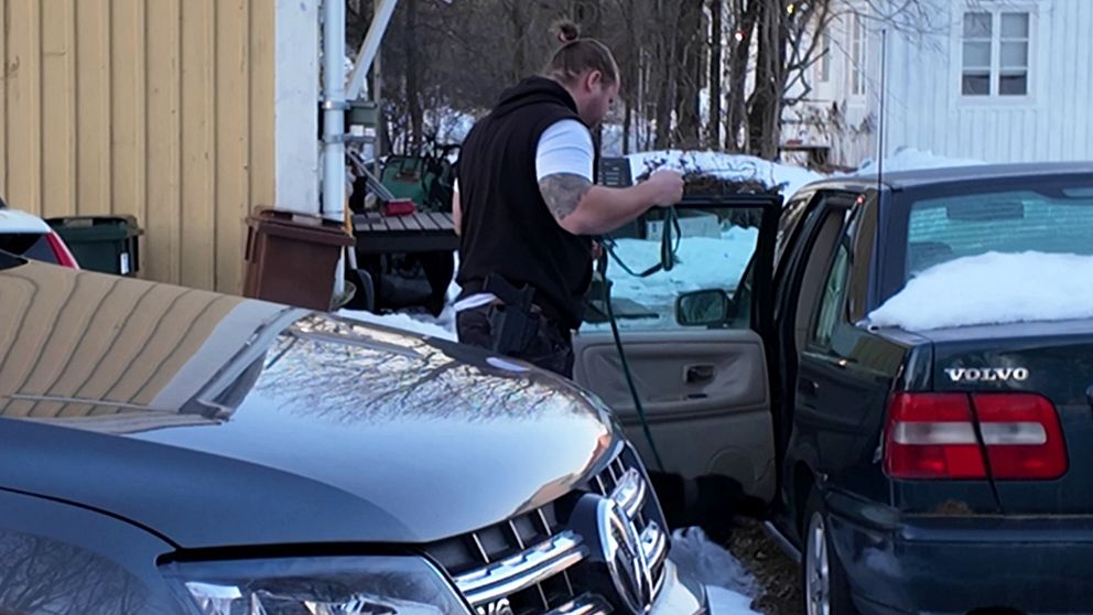 En polis står vid en bil som har en öppen dörr med en hund i koppel, I bakgrunden syns en byggnad och en annan bil.