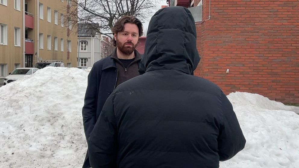 En anonym person som har på sig en svart jacka med luva. Mittemot står en reporter – en man med brunt hår