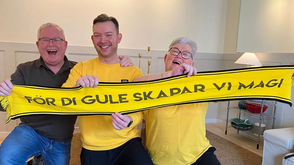 Tre personer sitter bredvid varandra och håller upp en gul halsduk med texten ”FÖR DI GULE SKAPAR VI MAGI”. De två personerna på sidorna har på sig gula tröjor.