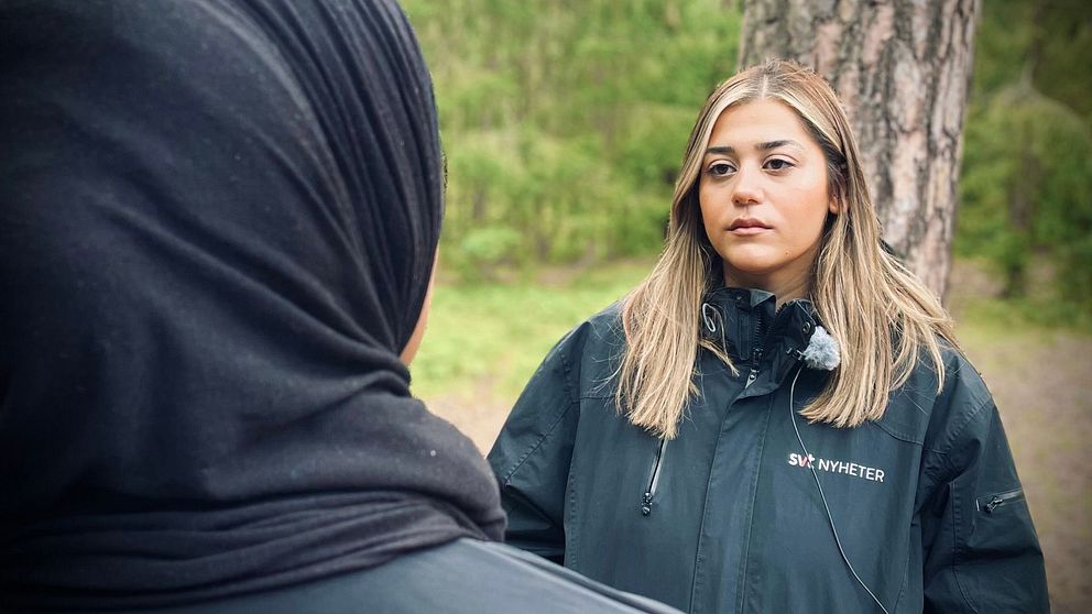 Två personer står vid en trädstam i en grönskande miljö, med fokus på en intervju. Den ena personen bär en svart klädsel, medan den andra har långt, ljust hår och en svart jacka med en logotyp på bröstet. Den omgivande naturen ger en lugn bakgrund för samtalet.