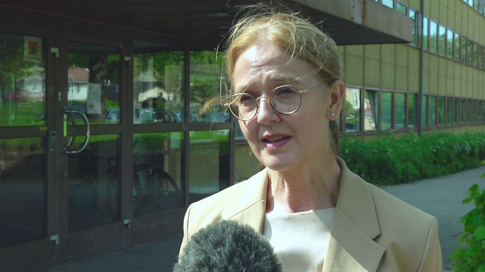 En kvinna i beige kavaj står framför en gul byggnad