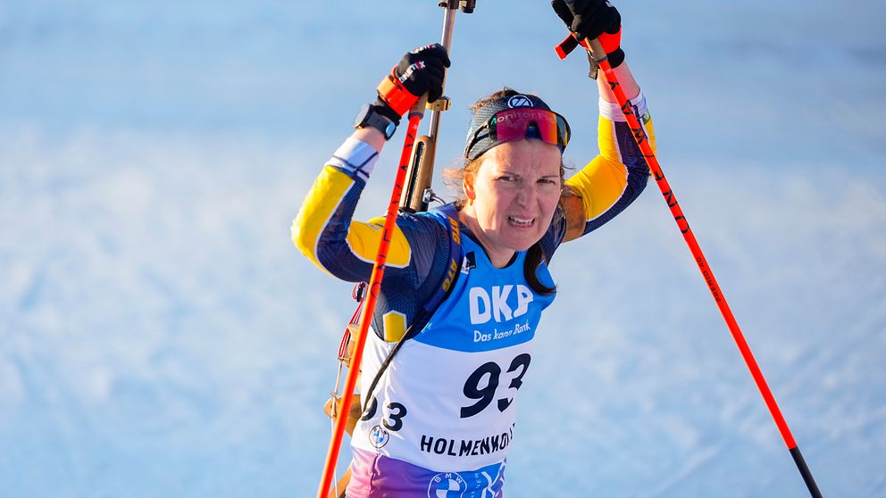 Skidskytt i tävlingsdräkt jublar med stavar i luften på snötäckt arena.