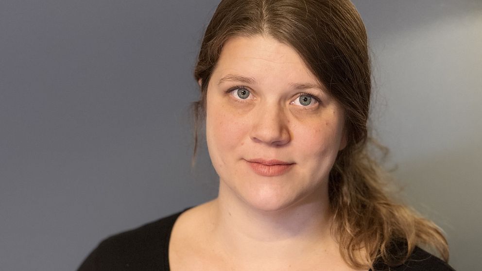 En kvinna med mörkt blont hår uppsatt i en låg hästsvans syns. Hon bär en svart tröja. Bakgrunden är en oskarp grå vägg.