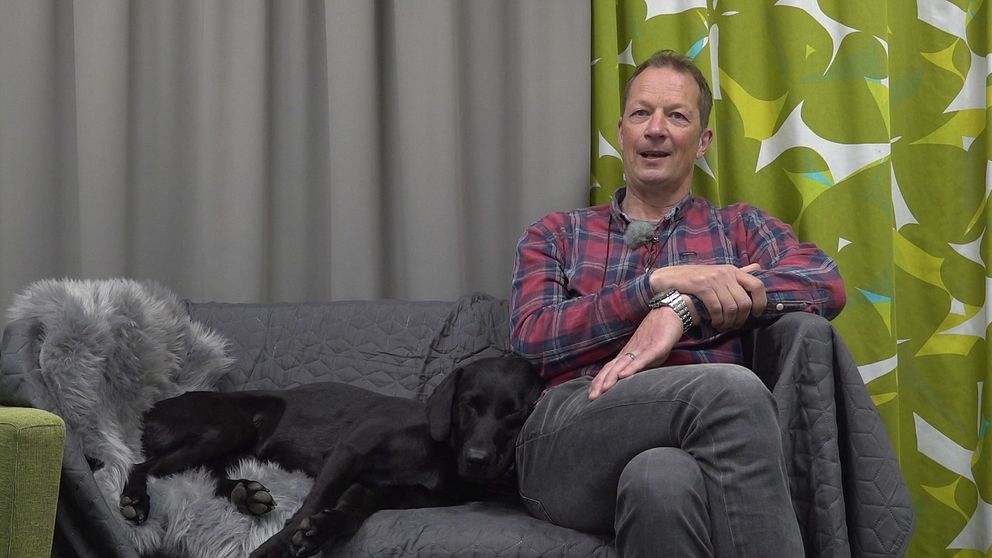 Den svarta jaktlabradoren Doffe med hundförare Karl Persson sitter i en soffa.