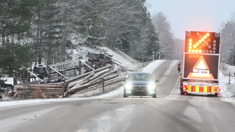 Ett vinterväglag där en varningsskylt med texten ”Bärgning” och blinkande ljuspilar står parkerad på landsvägen, medan en personbil närmar sig. På sidan av vägen ligger en vält lastbil med timmer utspritt, omgiven av snötäckta träd.