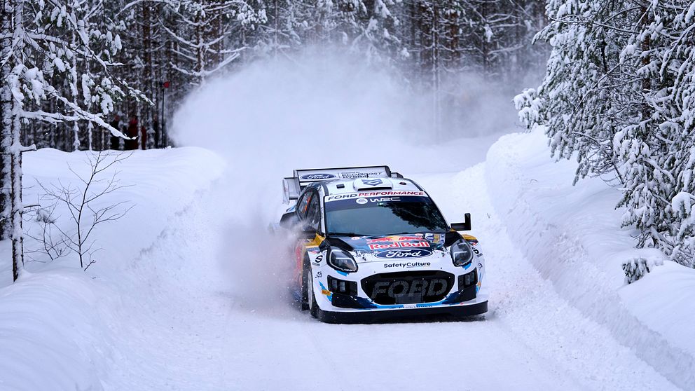 En rallybil kör på en snötäckt väg omgiven av tallar, med snö som sprutar upp bakom bilen.