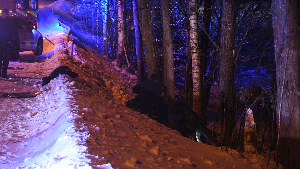 blåljus på snöig väg, en person arbetar vid en bil som ligger i det branta diket bland träd