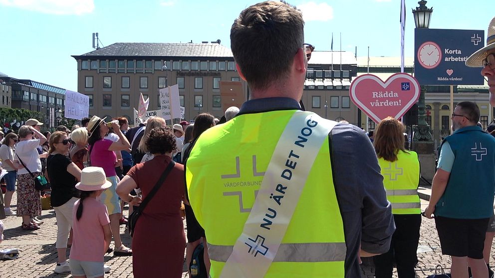 Flera personer strejkar på ett torg. En man med gul väst från Vårdförbundet har ett budskap på ryggen med texten ”nu är det nog”.
