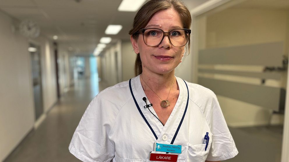 En läkare iklädd vit labbrock med namnskylt i en ljus sjukhusmiljö, stående framför en lång korridor.