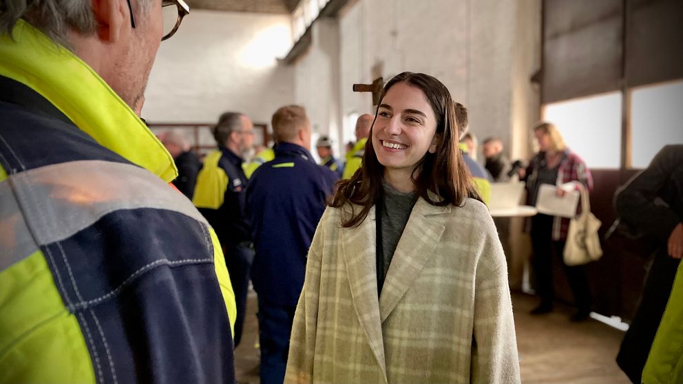 Personer i arbetskläder samtalar i en industribyggnad. Fler personer i bakgrunden.