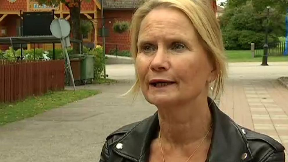 Kvinna i svart läderjacka står på en parkväg, omgiven av grönska och byggnader i bakgrunden. Det är en klar dag och en parkstruktur syns i avståndet.