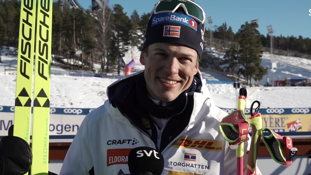 En skidtävlande person står med en mikrofonskylt från SVT i förgrunden, omgiven av snö och träd i bakgrunden. Personen håller skidor och stavar, och bär skidkläder med sponsormärken.