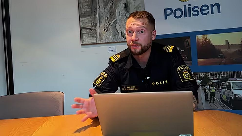 En polis, klädd i uniform, sitter vid ett bord framför en laptop. Bakgrunden visar en vägg med en stor skylt med texten ”Polisen” och en bild som relaterar till polisarbetet. Polisen gestikulerar med handen, vilket antyder att han diskuterar något viktigt.