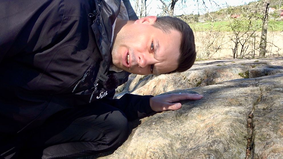Man i mörk jacka lutar sig ner och betraktar en klippa utomhus.