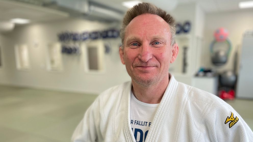 En person i judodräkt står i en träningslokal med pokaler i bakgrunden.