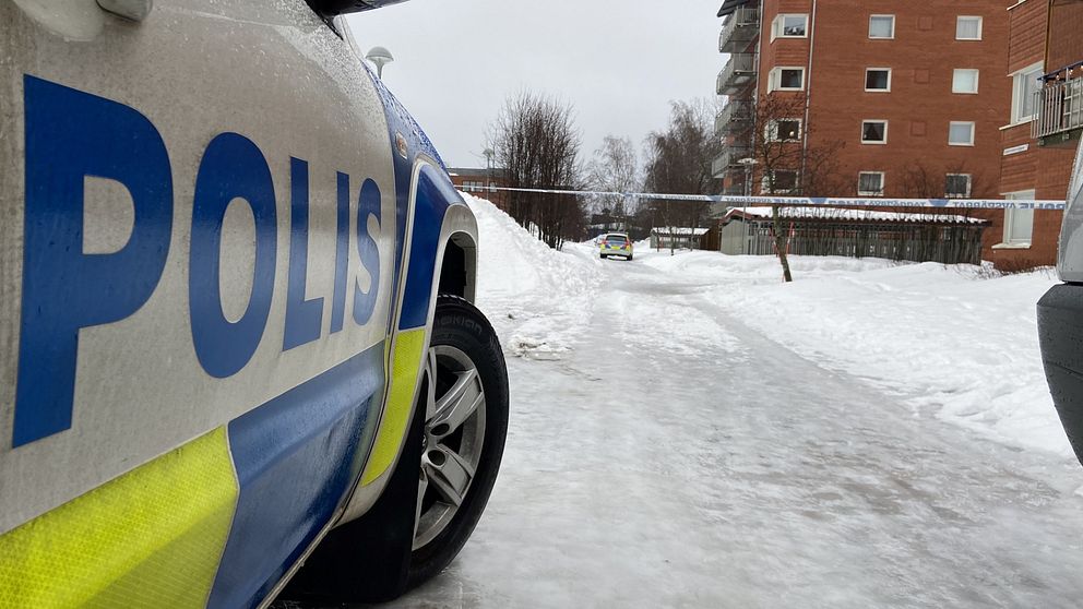 En polisbil står parkerad på en isig väg i ett bostadsområde. I bakgrunden syns flerbostadshus och snötäckta träd, medan en annan polisbil står längre ner längs vägen.