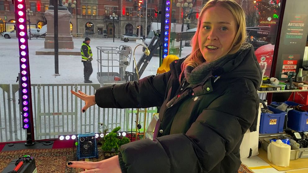 En person står i Musikhjälpens bur och håller ut armarna. Bakom personen syns en man i uniform som arbetar i området, samt en lyftplattform och en stängselstruktur. Den omgivande staden har lampor som lyser och skapar en livlig atmosfär.