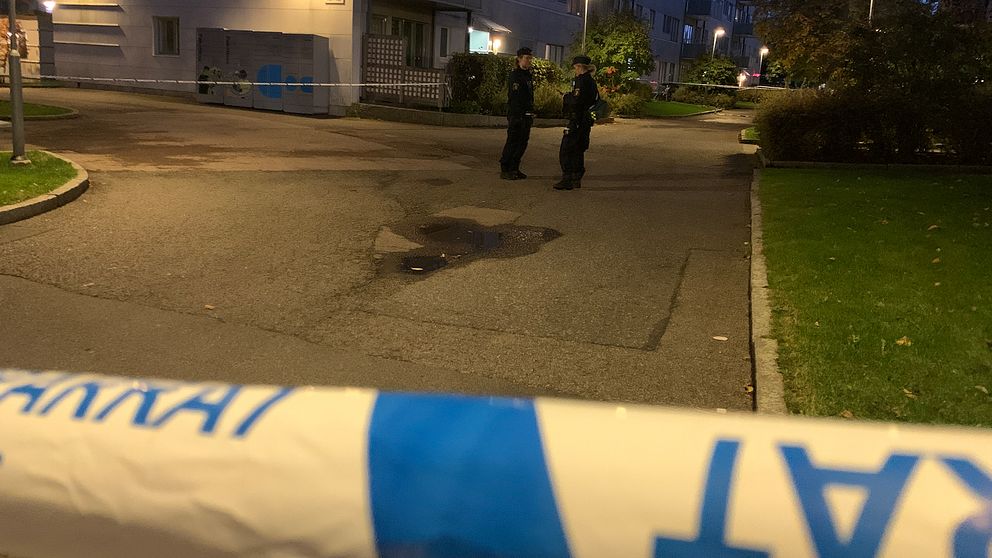 ALT-text: Två poliser står i en mörk bostadsområde där avspärrningar med polisband syns i förgrunden. En eventuell brottsplats är markerad i gatan.