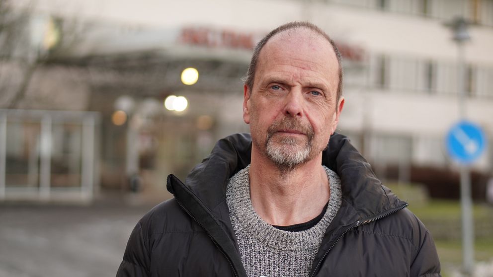 En person i en svart jacka och en grå tröja står framför en byggnad med en skylt. Personen bär ett namnskylt på bröstet. I bakgrunden syns en parkeringsskylt och träd.