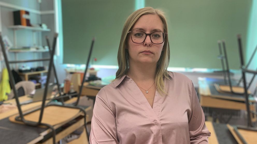 Kvinna i en rosa skjorta står framför en klassrumsmiljö med bord och stolar i bakgrunden. Ljusa fönster ger naturligt ljus till rummet. Lärare, lärarbehörighet, skola, varför få behöriga lärare?