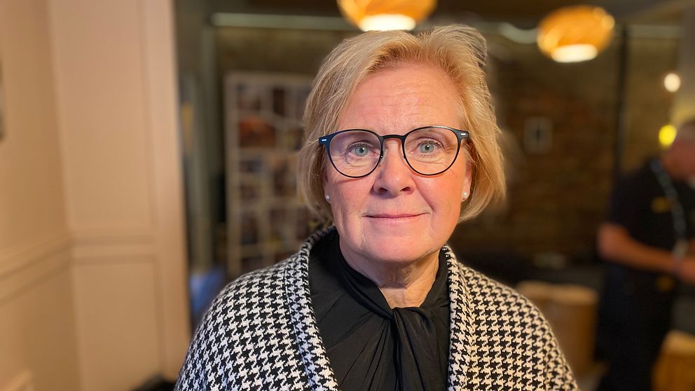 En kvinna står i en inomhusmiljö, iförd en svart och vit rutmönstrad kofta över en svart blus. I bakgrunden syns mjukt belysta lampor och en otydlig silhuett av en annan person. Atmosfären är avslappnad och hemtrevlig.