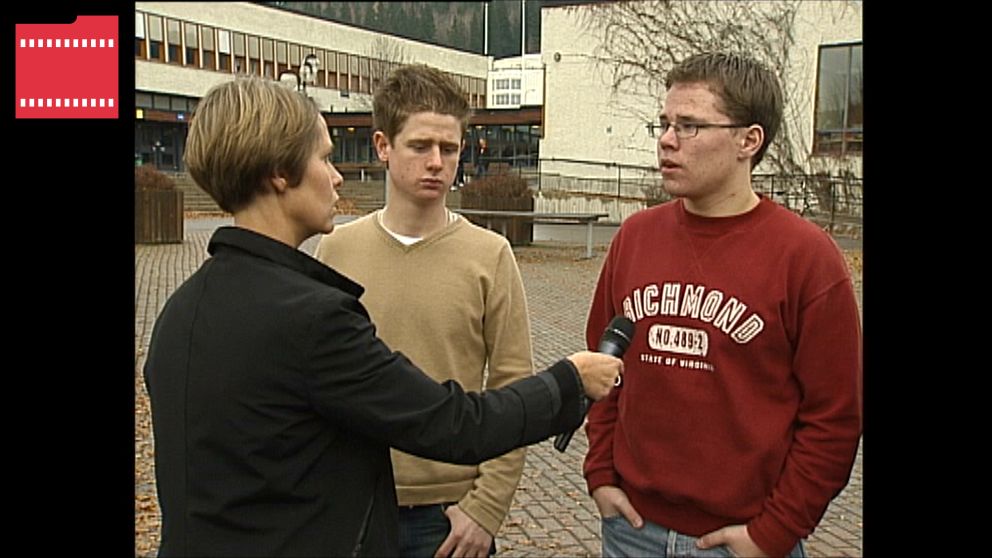 En journalist intervjuar två unga män framför en byggnad med parkeringsplatser och träd omkring. Den ena mannen bär en beige tröja och den andra en röd tröja med texten ”Richmond”.