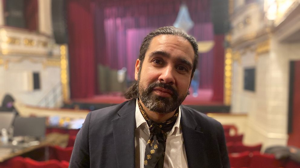 En person står i en teater med röda stolar i bakgrunden. Ridån är öppen och en scen med scenografi syns svagt belyst längst bak i lokalen.