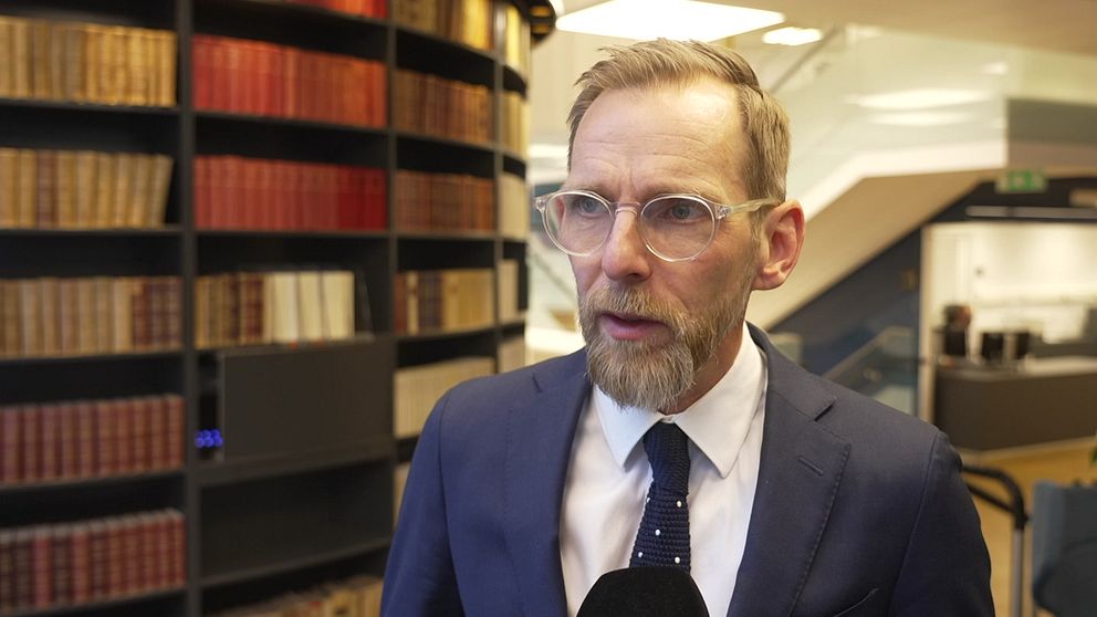 En man klädd i en mörk kostym med slips står framför en bokhylla fylld med böcker i en modern inomhusmiljö. I bakgrunden syns mörka och ljusa ytor som skapar en professionell atmosfär.