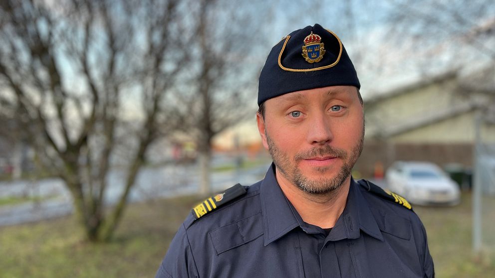 En man i uniform med en militärisk kap över huvudet står framför en suddig bakgrund med träd och byggnader. Han ser fokuserad ut och är klädd i en mörkblå skjorta med guldiga insignier på axlarna.