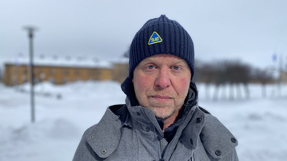 Slao:s vd Magnus Noppa, klädd i en grå vinterjacka och mörk mössa står utomhus på en snöig plats, med en suddig byggnad och träd i bakgrunden.