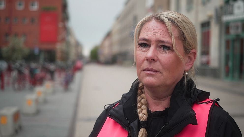 Kvinna med fläta och reflexväst står på en stadsgata med suddig bakgrund.